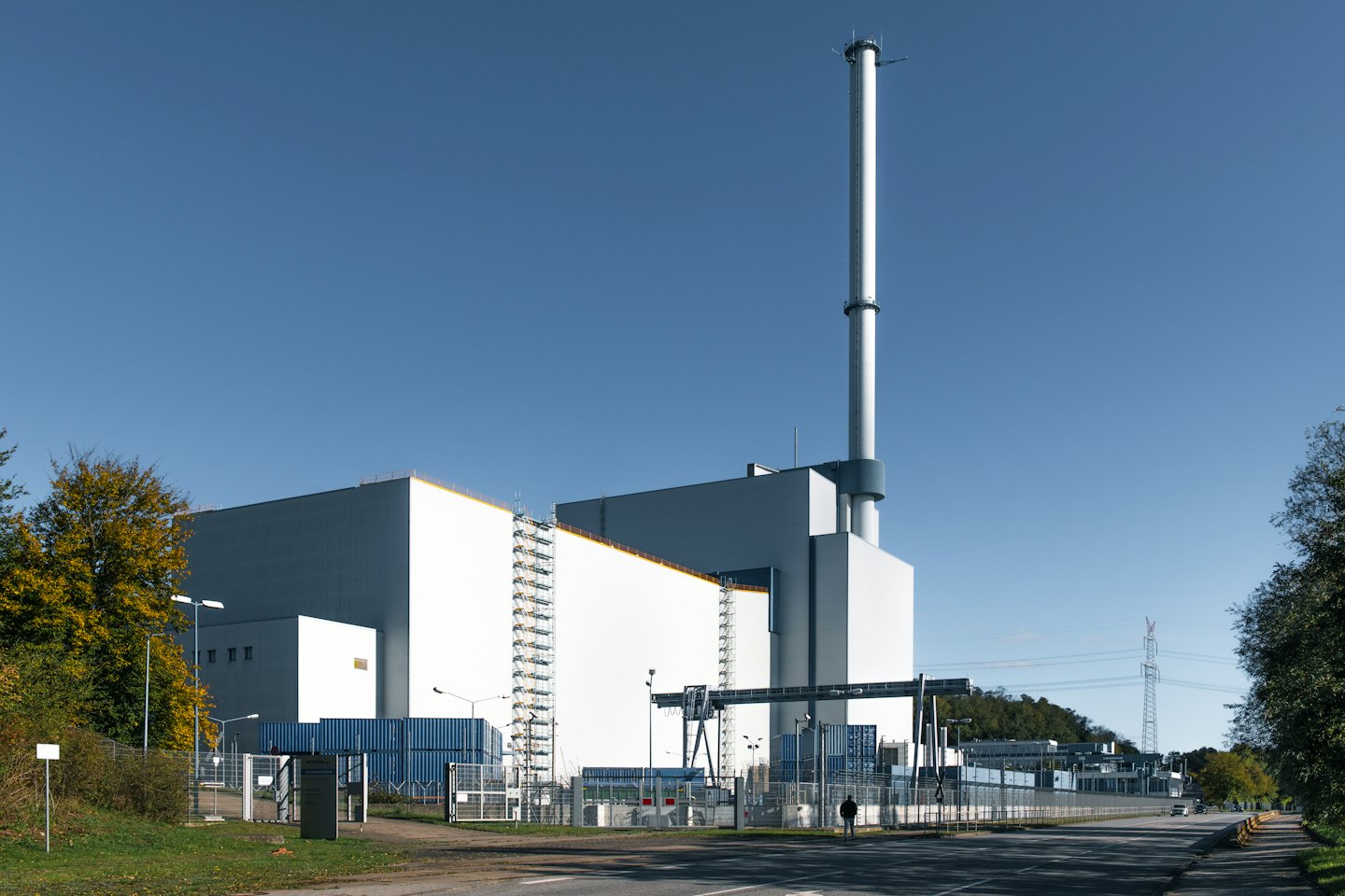 Person walking past a modern industrial facility with autumn trees.