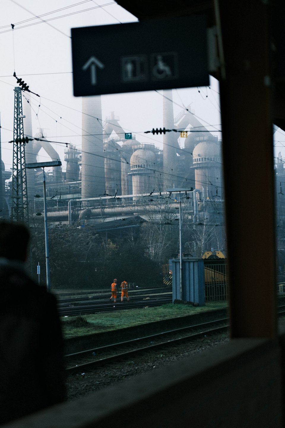 Two workers in orange vests walk between train tracks towards a factory.
